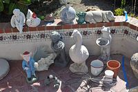 Photo showing a variety of garden statues including a blue-suited figure, small frog, white goose statues, ceramic pots, and a small hippopotamus statue lying down.