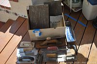 Wide view of the lot showing a cardboard box containing several wooden floats and metal trowels with wooden handles, two wooden floats outside the box plus a metal hammer and several additional metal trowels on the floor