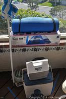 Photo of dome tent box stacked with blue rolled sleeping pad, MiniMate Ultra cooler on a larger cooler, with blue and white umbrella to the left, outdoor balcony setting with greenery and suburban street view.