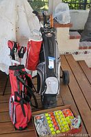 Full lot with red Ping golf stand bag filled with clubs, black and gray cart bag, and box with golf balls, glove, and tees on wood deck.