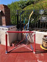Hockey net and four hockey sticks arranged against a wall outdoors, showing overall lot contents and condition.