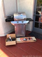 Char Broil Commercial Infrared Grill with wheels, black grill cover, Fire Magic motorized spit and box of accessories arranged in front.