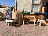 Wide view of lot outdoors on paver surface showing saw horse table, cinderblocks, trash cans, plywood, wooden panels, and other items