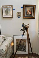 Vintage standing wine opener on wooden tripod with four pieces of framed wall art visible on wall and floor
