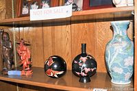 Shelf displaying the two wood statues - brown and reddish, two round black lacquered bowls with floral design, and blue floral vase.