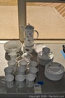 Full view of the entire fine china set arranged on surface, showing coffee pot on warmer, cups, plates, saucers, milk and sugar containers, bowl, and trays.