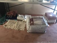 Overview of folded blankets, linens, and packaged comforter on carpeted floor with folding table in background