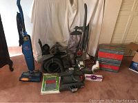 Wide view showing Kirby vacuum with cloth bag, Bissell Steam and Sweep blue upright vacuum, various attachments, boxed vacuum bags, and carpet shampoo bottle arranged on carpeted floor.