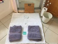 White bath mat laid flat with two folded light purple hand towels and bathroom scale behind.