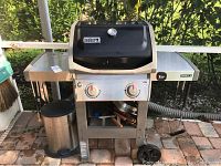 Weber Spirit II gas grill with side shelves and trashcan beside it, showing front view.