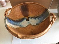 Top-down angled view of the pottery water bowl showing the swirl of blue glaze across the brown interior and one rope-style handle visible.