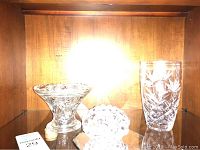 View of three items on a wooden shelf: tall glass vase, clear glass dish, and a glass frog figurine.