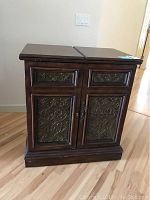 Front view of wood bar on wheels with ornate metal panels on drawer and door faces, showing the closed top with a split design.