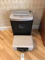 Fellows paper shredder standing behind the Sentry lock box on a wooden floor against a wallpapered wall.