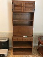 Full front view of the entire wooden secretary desk showing upper cabinet with double louvered doors, open middle shelf, fold-down desk surface, drawer section below, and open bottom shelf.