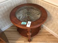 Full view of round table showing clear glass top with detailed floral relief in the center, wooden rim, and legs ending in claw feet.