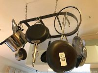 Photo showing multiple pots and pans hanging from a black metal rack suspended by chains. Includes frying pans and saucepans.