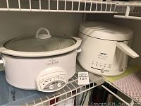 Photo showing the Rival Crock-Pot slow cooker on the left with glass lid and T-Fal MaxiFry deep fryer on the right side on a shelf.