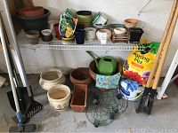 Wide view of garden tools and pots on floor and shelf including rakes, shovels, watering can, plant support frames, and bags of potting mix.