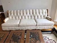 Front view of a light beige upholstered sofa with tufted backrest, wooden frame with curved armrests and carved legs, placed indoors with coffee table in front.