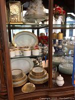 Display cabinet with multiple shelves showing the Royal Gallery gold accent china service, glass cake server on top shelf, Kundo clock, Polish ceramic candlesticks, and other glassware and decorations.