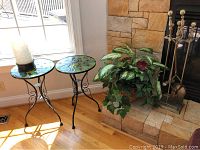 Photo of two peacock feather pattern glass top plant stands beside a window with sunlight, the top of one holds the flameless candle holder with candle, stone fireplace and faux plant visible.