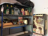 Metal shelving unit with two bottles of Ortho Weed B Gon Lawn Weed Killer spray, other garden items, Black & Decker circular saw box, and illuminated standing reindeer box.