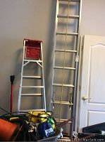 Image showing both ladders against wall alongside tools and other items, includes red platform step ladder and tall extension ladder with yellow rope.