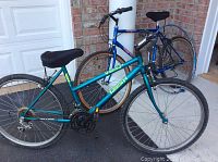 Two adult bicycles, one turquoise Triumph Vertical DX 15-speed mountain bike and one blue Venture ATB Canyon Runner 18-speed mountain bike, parked on pavement outside a brick building.