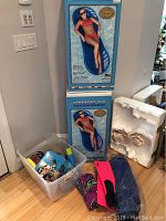 Two boxed Emerald Isle pool floats stacked, plastic bin with goggles, flippers next to box.