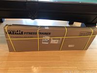 Sealed large rectangular cardboard box with 'Tony Little's Gazelle Fitness Trainer' branding and shipping address, secured with yellow straps.