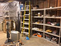 Wide view showing four plastic shelving units filled with miscellaneous items in a garage setting; includes a yellow ladder and some gold-colored framed items nearby. Shelves are beige/tan plastic and appear sturdy.
