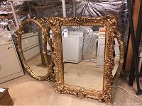 Two ornate gold framed wall mirrors, one rectangular and one oval, shown in basement environment.