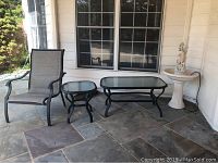 Photo showing outdoor furniture set arranged on patio: one mesh fabric chair with metal frame, small square glass-top end table, larger rectangular glass-top coffee table, and electric bird bath with pedestal base and decorative top.