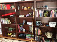 Wide shot of three wooden shelving units filled with books, toys, board games, and decorative items including a green dinosaur figurine, Monopoly game box, and various books mainly study guides and educational.
