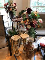 Photo showing two gold and white decorative pillar stands, large decorated ceramic vase, some artificial flowers, and part of the brass and wooden plant stands grouped near a window.