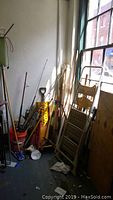 Multiple yard and car tools including fishing rods, rakes, shovels, and brooms placed near a window on a floor