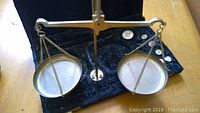 Brass balance scale mounted in black carrying case with two round pans suspended by chains.