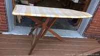 Antique wooden ironing board with yellow and white striped fabric cover shown from side view.