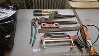 Photo showing assortment of hand tools including wrenches, caulking guns, levels, hacksaws, and bolt cutter on tabletop