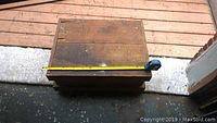 Top-down view of the closed wooden chest on a porch, showing weathered wood with a measuring tape indicating size approximately 26.5" long.
