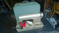 Kenmore sewing machine in metallic light gold with carrying case in light green behind it. Machine is on a stand with foot pedal and power cord attached, spool of white thread loaded on the machine.