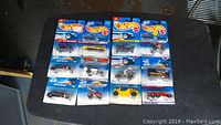 Top-down photo showing 12 Hot Wheels cars in original blue packaging arranged on a black surface. All cars sealed in blister packs.
