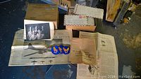 Box with a large stack of letters, some loose envelopes, AAC patches, and folded newspaper pages visible on a dark floor.