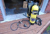 Photo of vintage yellow Aqua Lung SCUBA tank standing upright with regulator, pressure gauge and hoses laid out on wooden deck outside a garage.