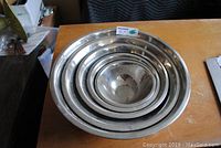Five nested stainless steel mixing bowls on a wooden surface, showing the full set with visible usage marks.