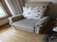 Upholstered beige recliner chair with a decorative leaf pattern pillow placed against the backrest. The chair is positioned near a window and sits on a wooden floor.