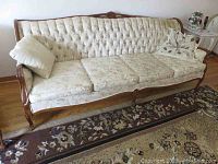 Front view of carved wood and upholstered beige sofa with three seat cushions and two pillows on a floral patterned rug.