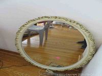 Full view of an oval wall mirror with an ornate cream-colored frame placed on a hardwood floor, showing the reflective glass and part of a chair reflected.
