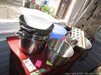Photo of stacked stainless steel pots, non-stick frying pan, white plastic colander, metal mixing bowls, and flatware container on a red table by a window.
